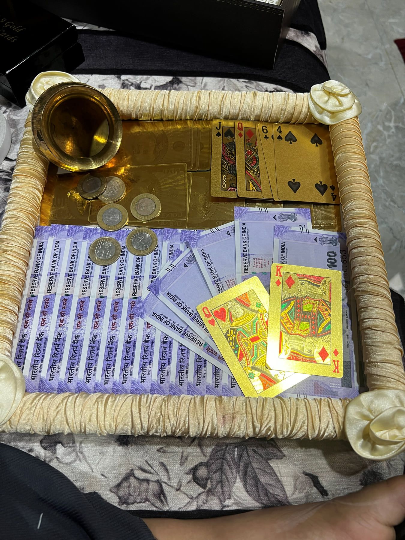 Gold shagun tray with brass kalash, coins, playing cards and currency notes framed in cream rope