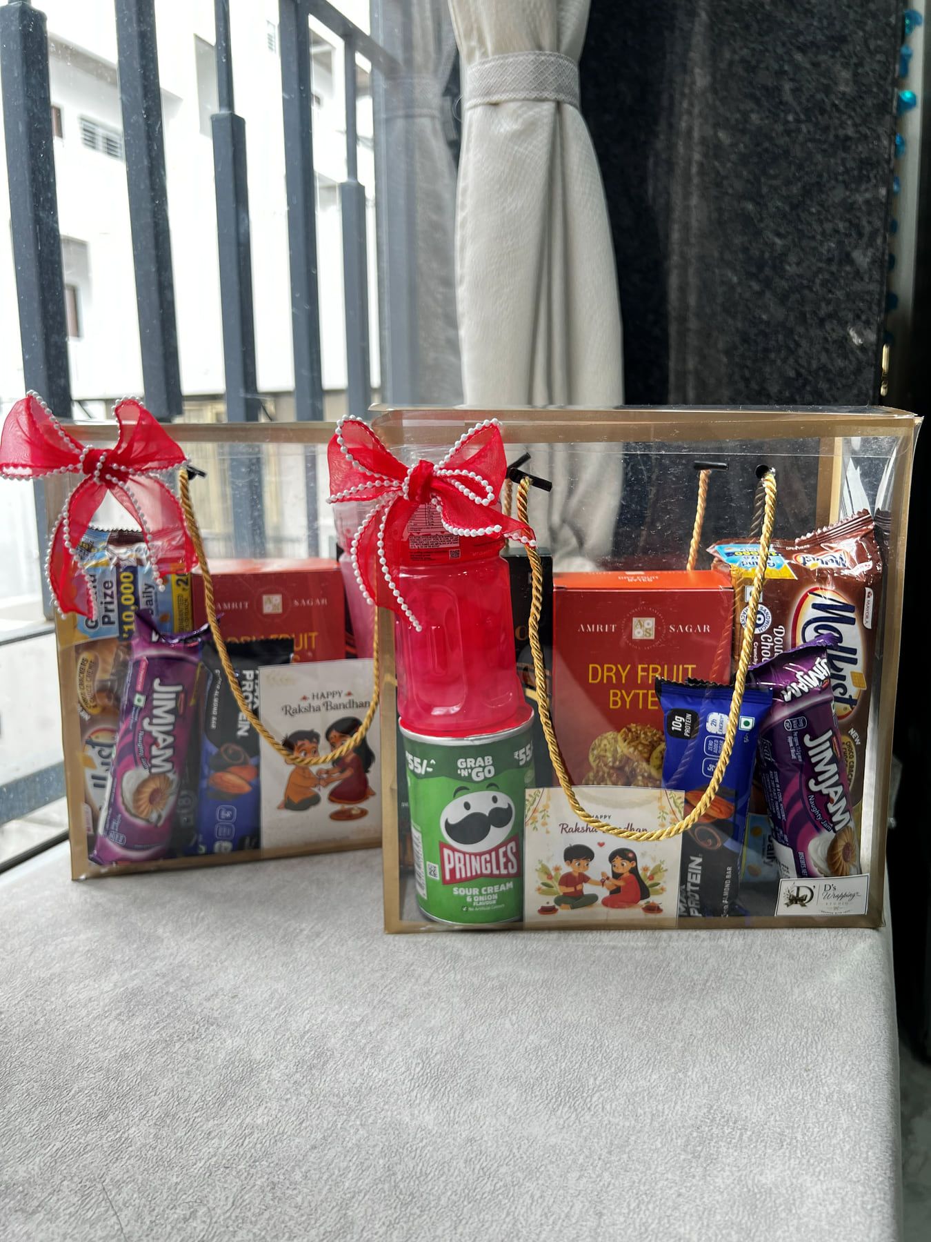 Two clear acrylic gift boxes with red satin bows filled with Pringles, chocolates, dry fruit bites and a rakhi card